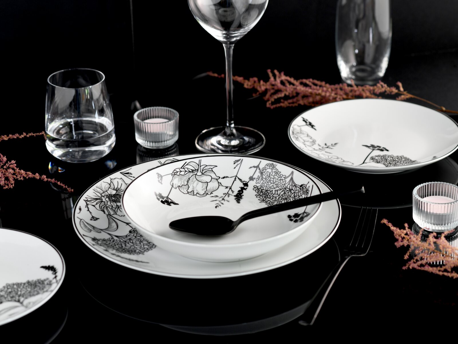 Elegantes weißes Porzellan-Tafelservice mit schwarzem floralen Dekor, Teller und Schale mit Löffel auf schwarzem Tisch, umgeben von Gläsern und Teelichtern, fotografiert aus leicht erhöhter schräger Perspektive