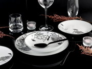 Elegantes weißes Porzellan-Tafelservice mit schwarzem floralen Dekor, Teller und Schale mit Löffel auf schwarzem Tisch, umgeben von Gläsern und Teelichtern, fotografiert aus leicht erhöhter schräger Perspektive