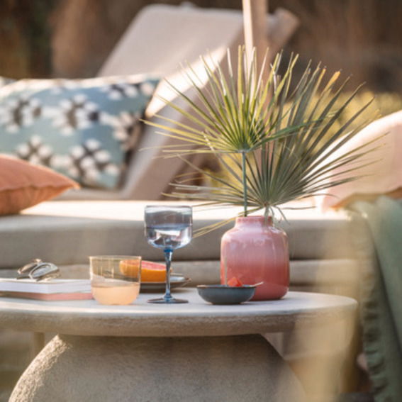 Like by Villeroy & Boch Weinkelch-2tlg. LIKE ICE Ein stilvoll gedeckter Tisch im Freien mit einem blauen Weinkelch aus dem Set 'Like Ice', umgeben von einem rosa Keramikgefäß mit Palmenblättern, einem weiteren Glas und einer Schale. Die Perspektive zeigt eine gemütliche Outdoor-Lounge-Atmosphäre.