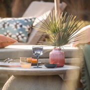 Ein stilvoll gedeckter Tisch im Freien mit einem blauen Weinkelch aus dem Set 'Like Ice', umgeben von einem rosa Keramikgefäß mit Palmenblättern, einem weiteren Glas und einer Schale. Die Perspektive zeigt eine gemütliche Outdoor-Lounge-Atmosphäre.