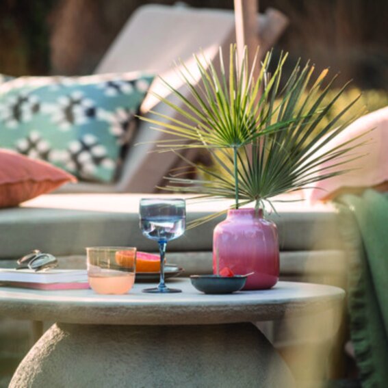 Like by Villeroy & Boch Weinkelch-2tlg. LIKE ICE Ein stilvoll gedeckter Tisch im Freien mit einem blauen Weinkelch aus dem Set 'Like Ice', umgeben von einem rosa Keramikgefäß mit Palmenblättern, einem weiteren Glas und einer Schale. Die Perspektive zeigt eine gemütliche Outdoor-Lounge-Atmosphäre.