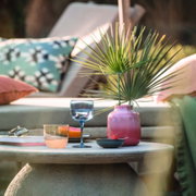Ein stilvoll gedeckter Tisch im Freien mit einem blauen Weinkelch aus dem Set 'Like Ice', umgeben von einem rosa Keramikgefäß mit Palmenblättern, einem weiteren Glas und einer Schale. Die Perspektive zeigt eine gemütliche Outdoor-Lounge-Atmosphäre.