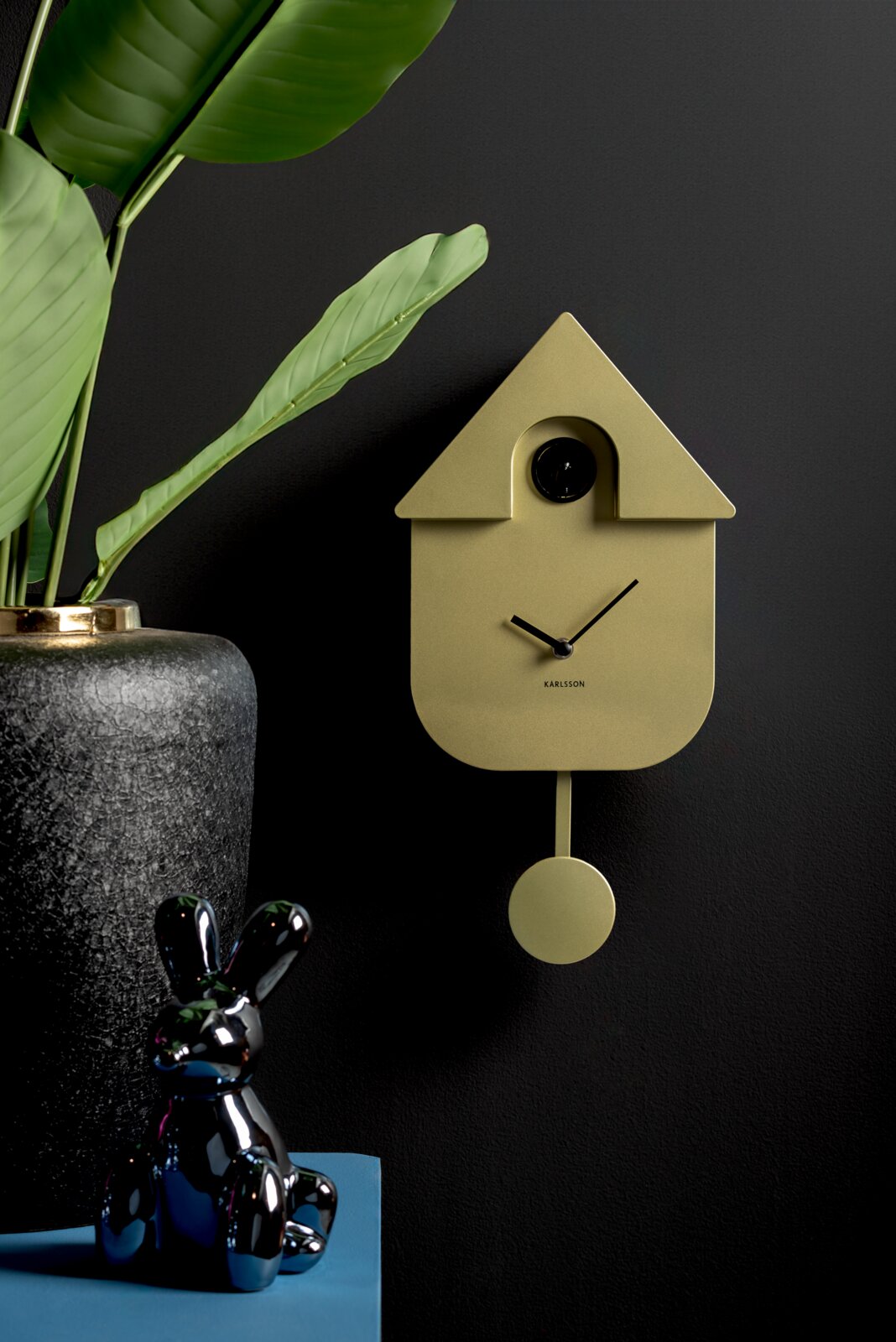 KARLSSON Wanduhr MODERN CUCKOO Moderne Wanduhr in Form eines Kuckucksuhrenhauses mit Pendel, seitliche Perspektive, neben einer Pflanze und einer dekorativen Figur.