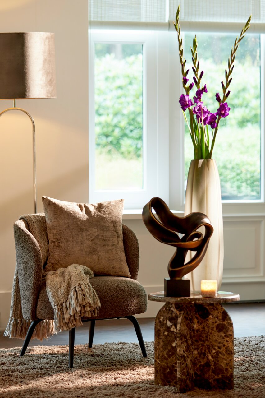 Light&Living Ornament KAJAN Modernes Wohnzimmer mit einem beigen Sessel, dekoriert mit einem Kissen und einer Decke, daneben ein kleiner Tisch mit einer abstrakten Skulptur und einer Kerze, im Hintergrund eine Vase mit lila Blumen. Perspektive: seitlich.
