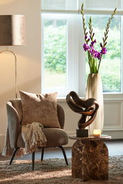 Modernes Wohnzimmer mit einem beigen Sessel, dekoriert mit einem Kissen und einer Decke, daneben ein kleiner Tisch mit einer abstrakten Skulptur und einer Kerze, im Hintergrund eine Vase mit lila Blumen. Perspektive: seitlich.