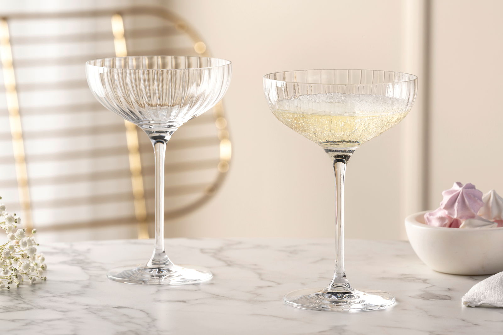 Zwei elegante Champagnerschalen POESIA aus Kristallglas, eine gefüllt mit Champagner, auf einem Marmortisch, seitliche Perspektive.