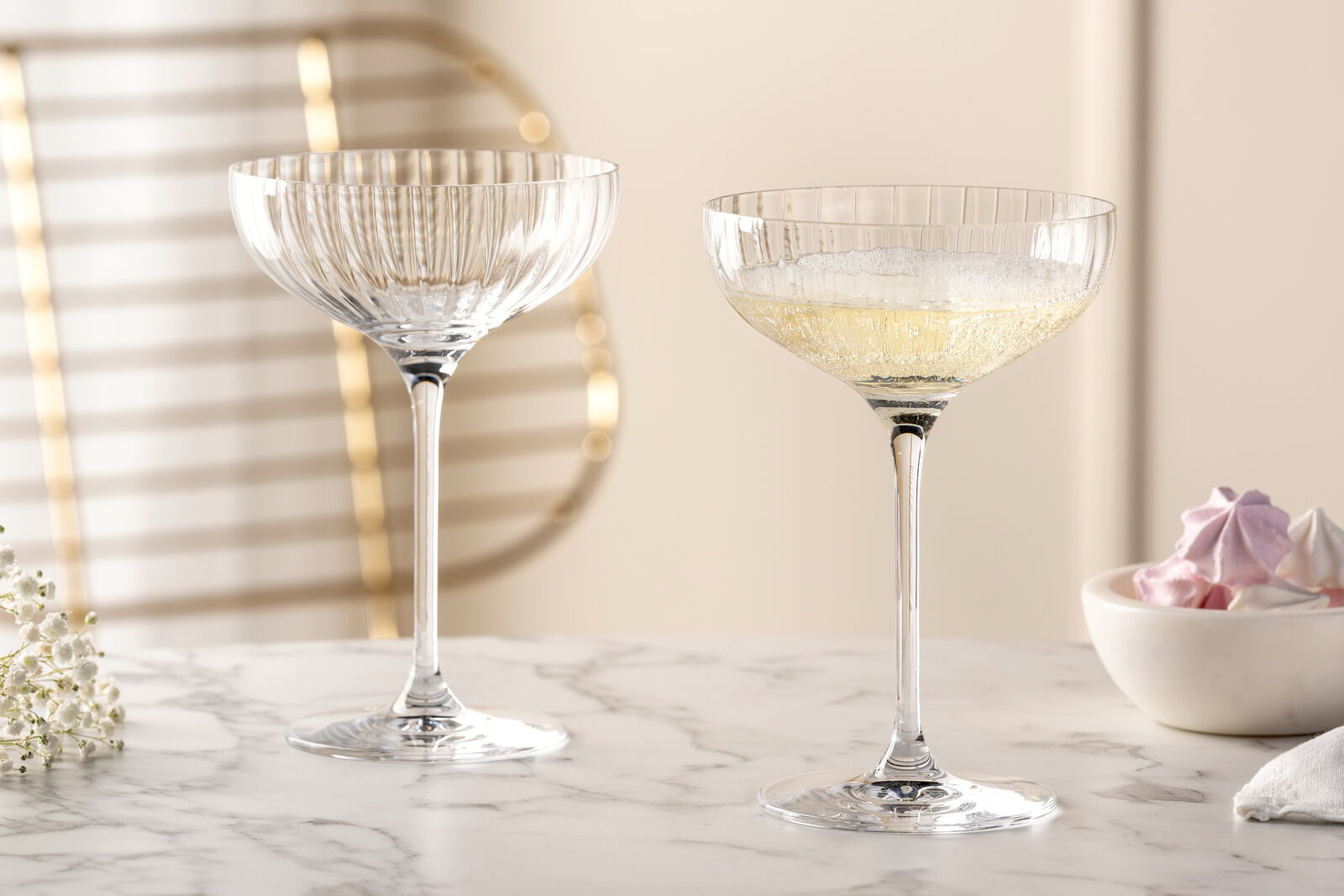 Zwei elegante Champagnerschalen POESIA aus Kristallglas, eine gefüllt mit Champagner, auf einem Marmortisch, seitliche Perspektive.