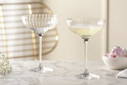 Twee elegante POESIA-champagneschalen van kristalglas, waarvan één gevuld met champagne, op een marmeren tafel, gezien vanuit de zijkant.