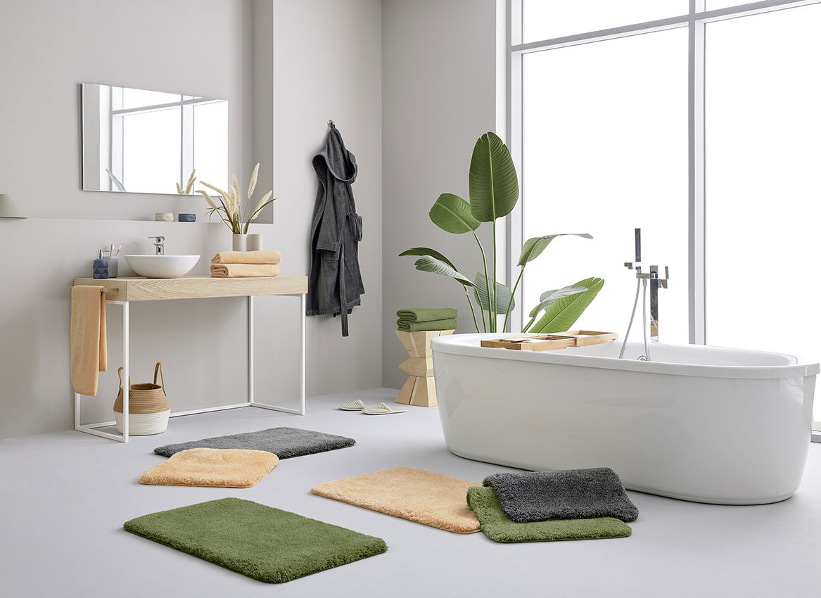 Moderne Badezimmeransicht mit mehreren Badteppichen in verschiedenen Farben auf dem Boden, neben einer freistehenden Badewanne und einem Waschtisch. Helle, minimalistische Einrichtung mit Pflanzen und Handtüchern. Perspektive von vorne.