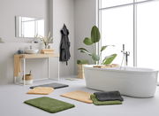 Moderne Badezimmeransicht mit mehreren Badteppichen in verschiedenen Farben auf dem Boden, neben einer freistehenden Badewanne und einem Waschtisch. Helle, minimalistische Einrichtung mit Pflanzen und Handtüchern. Perspektive von vorne.