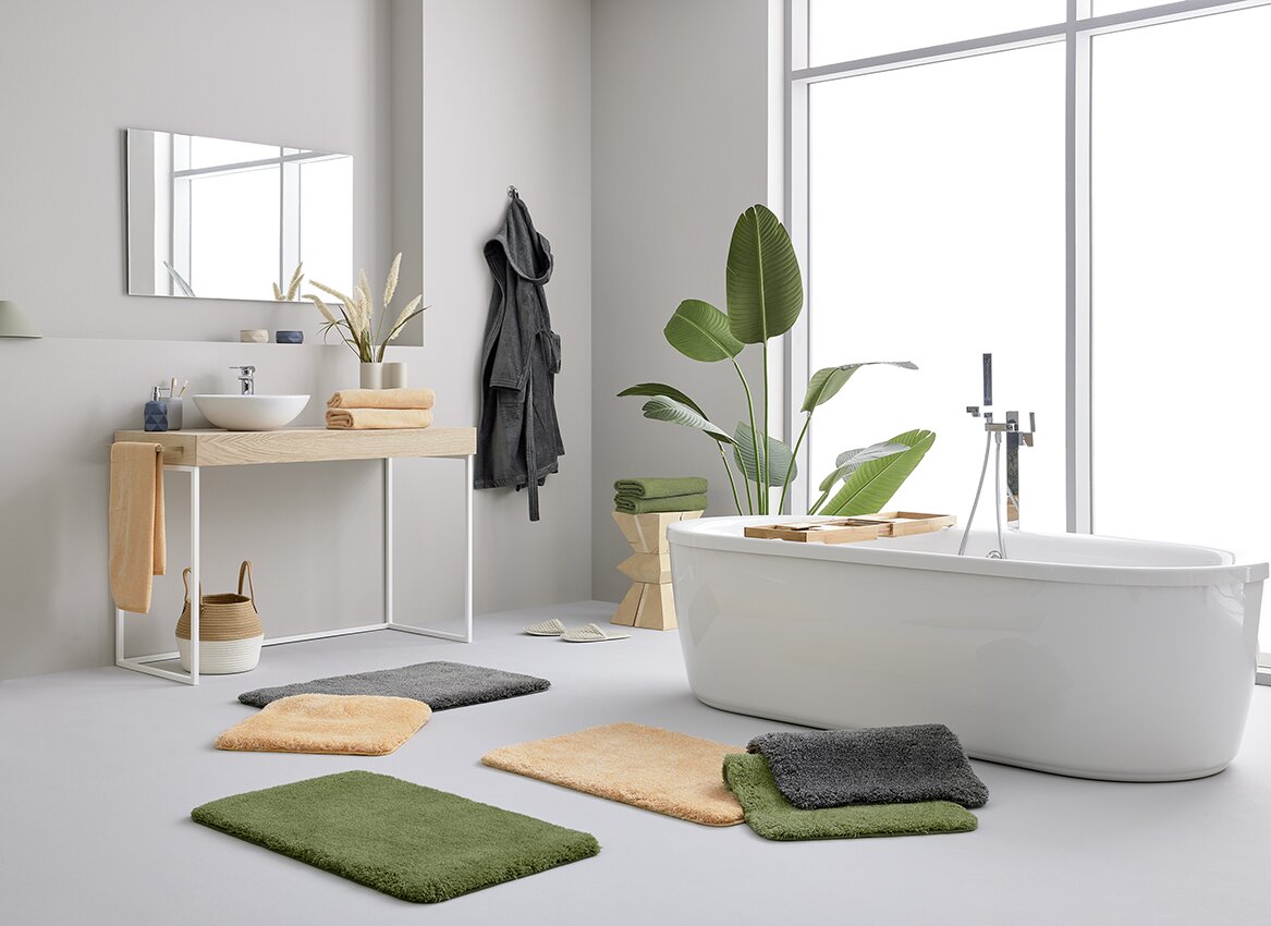 Een moderne badkamer met verschillende badkamermatten in diverse kleuren op de vloer, naast een vrijstaand bad en een wastafel. Een lichte, minimalistische inrichting met planten en handdoeken. Aanzicht van voren.
