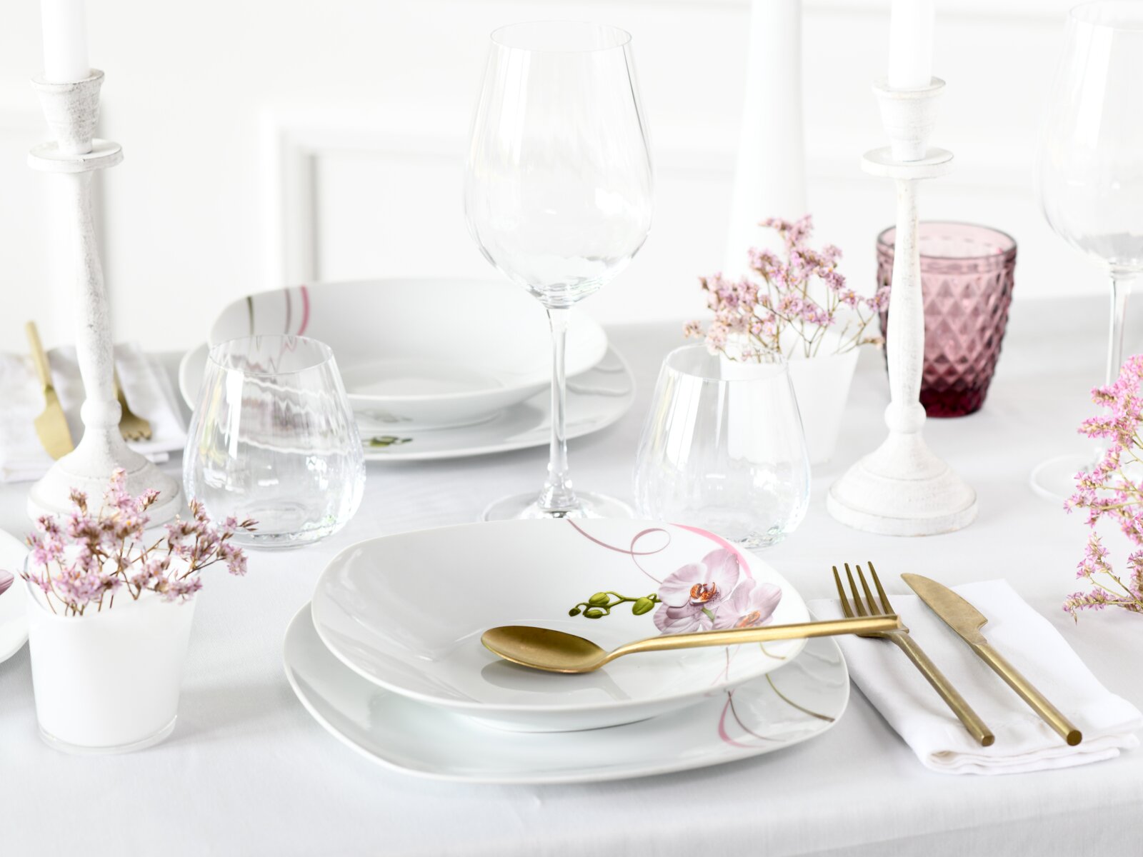 Elegantes weißes Tafelservice mit Orchideen-Motiv, goldfarbenem Besteck, Weingläsern und Kerzenständern auf festlich gedecktem Tisch, Perspektive: leicht erhöhte Frontansicht