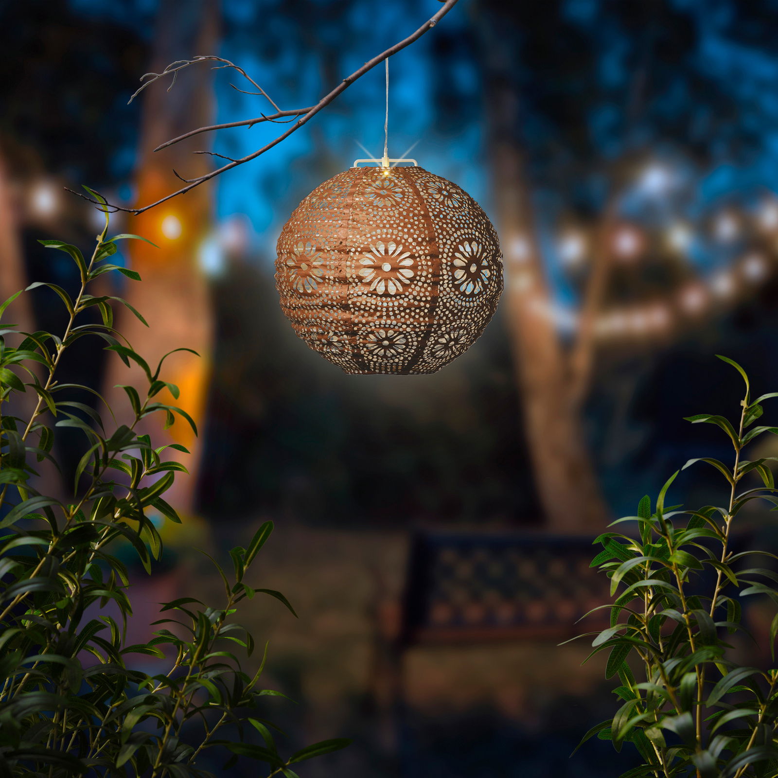 Solar Laterne mit filigranem Muster, hängend an einem Ast in einem stimmungsvoll beleuchteten Garten, seitliche Perspektive
