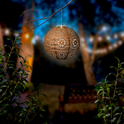 Solar Laterne mit filigranem Muster, hängend an einem Ast in einem stimmungsvoll beleuchteten Garten, seitliche Perspektive