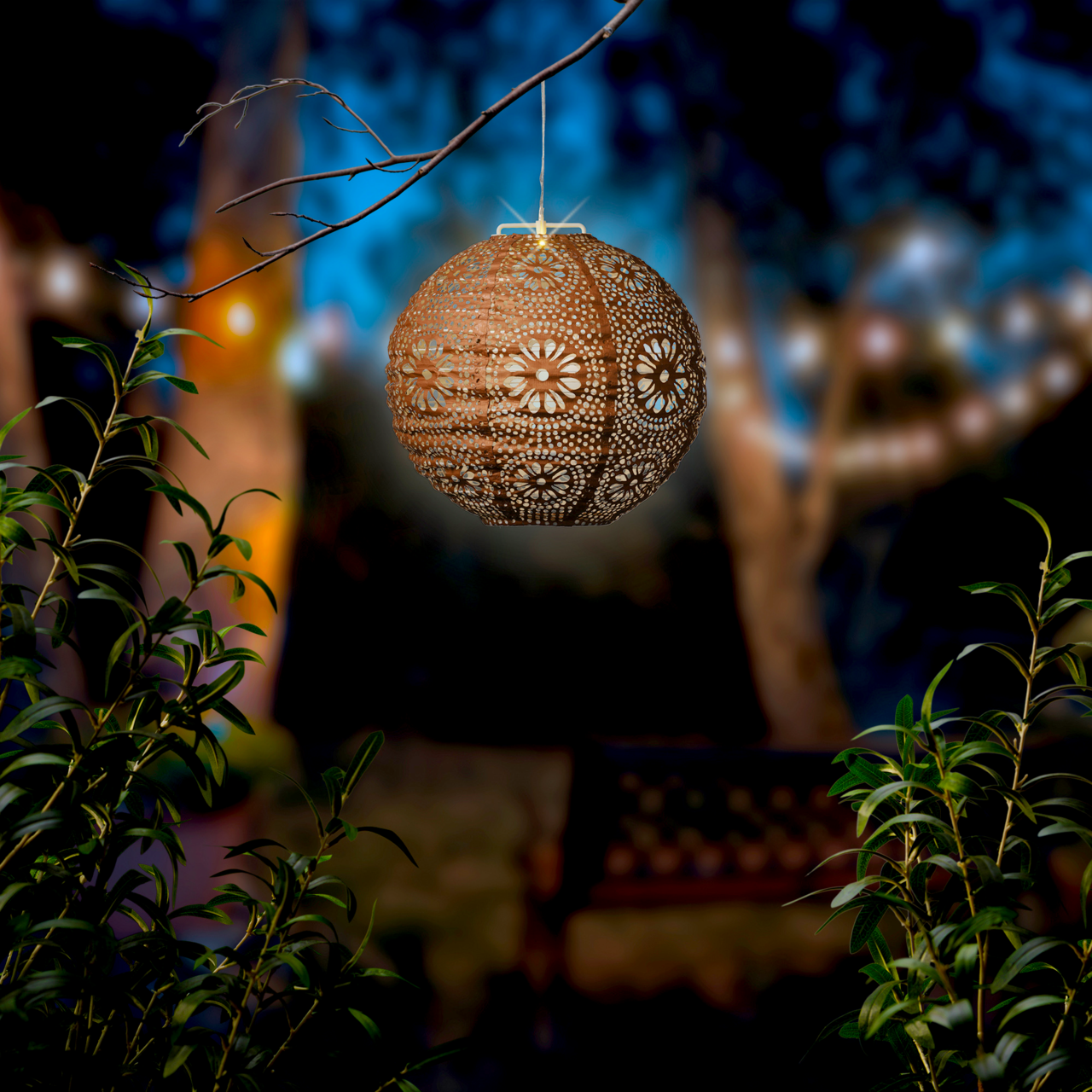 Solar Laterne mit filigranem Muster, hängend an einem Ast in einem stimmungsvoll beleuchteten Garten, seitliche Perspektive