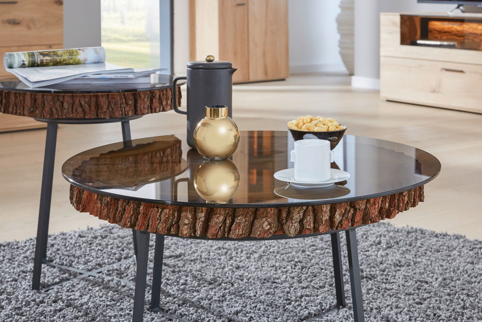 Runder Couchtisch mit Glasplatte und rustikalem Holzrand, seitliche Perspektive, dekoriert mit einer Tasse, einer goldenen Vase und Snacks.