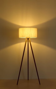Eingeschaltete Stehlampe mit beigem Stoffschirm und dreibeinigem Holzgestell, warmes Licht an der Wand, Frontansicht auf Augenhöhe.