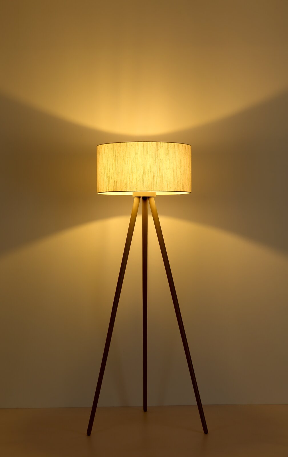Eingeschaltete Stehlampe mit beigem Stoffschirm und dreibeinigem Holzgestell, warmes Licht an der Wand, Frontansicht auf Augenhöhe.
