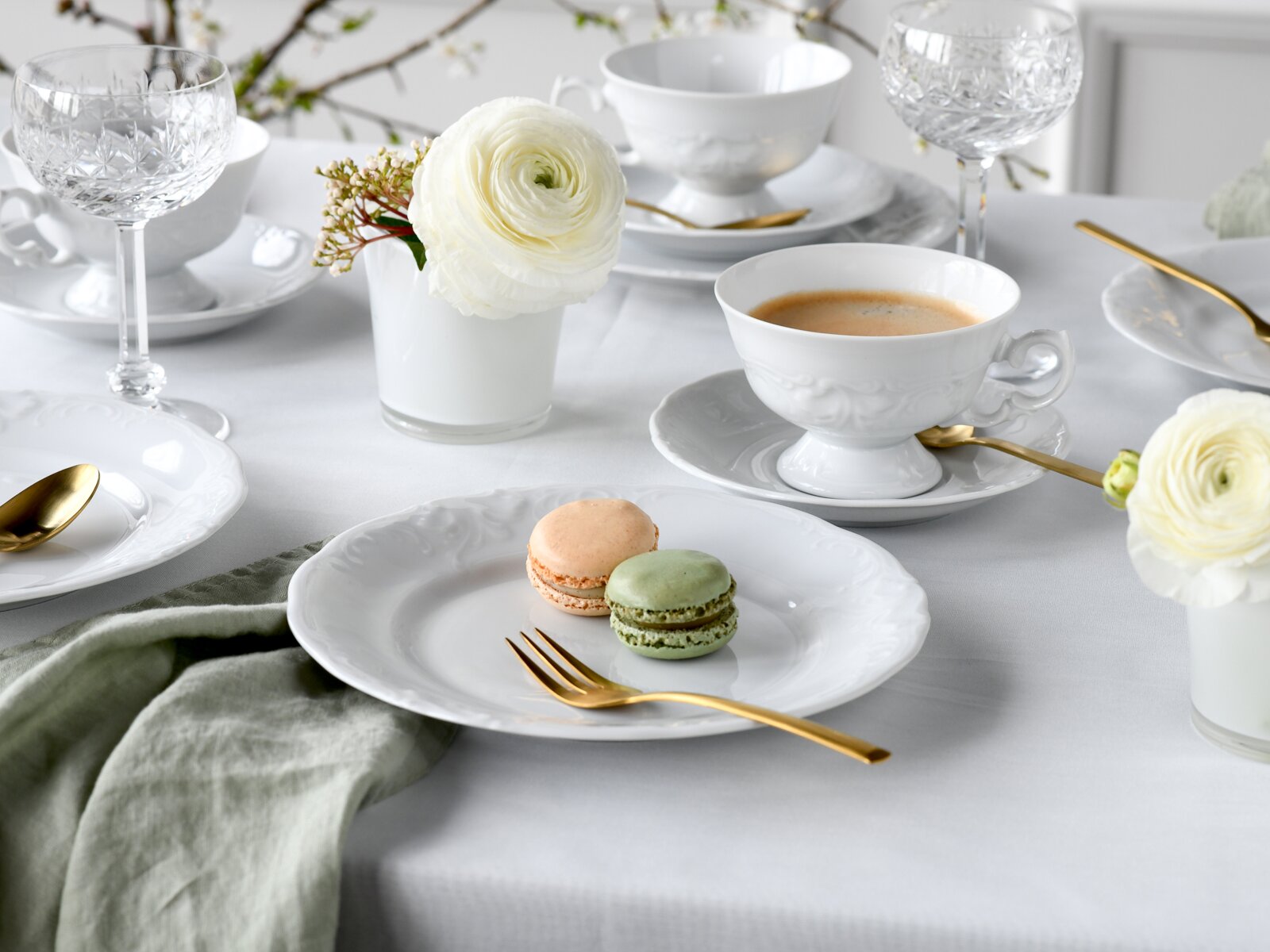 Elegantes weißes Porzellan-Kaffeegeschirr mit Reliefdekor, goldfarbenem Besteck und Kristallgläsern auf gedecktem Tisch, im Vordergrund Tasse mit Kaffee und Teller mit zwei Macarons, perspektivisch in leicht schräger Ansicht von vorne oben