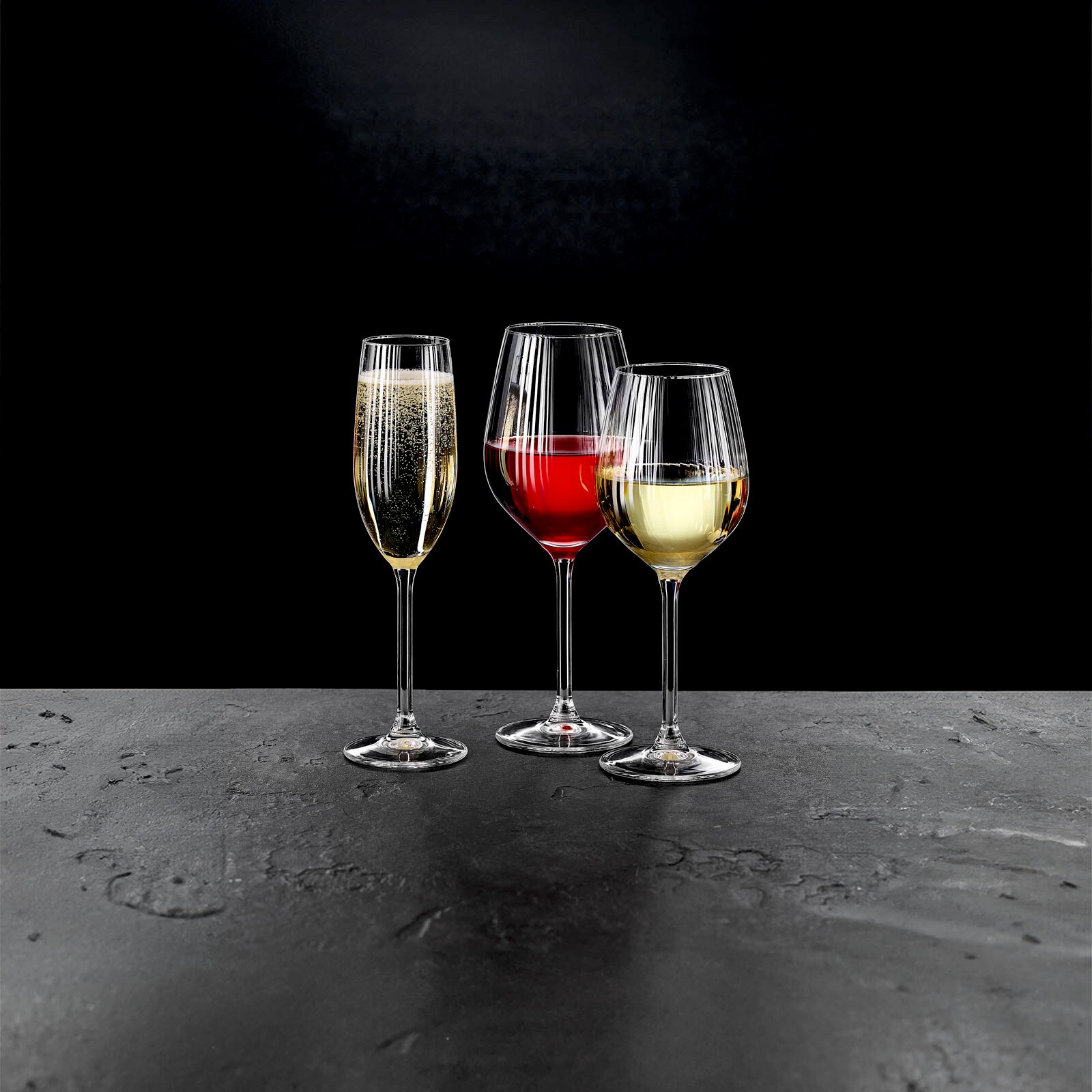 Drei Stielgläser mit Sekt, Rotwein und Weißwein auf einer dunklen Oberfläche, seitliche Perspektive.