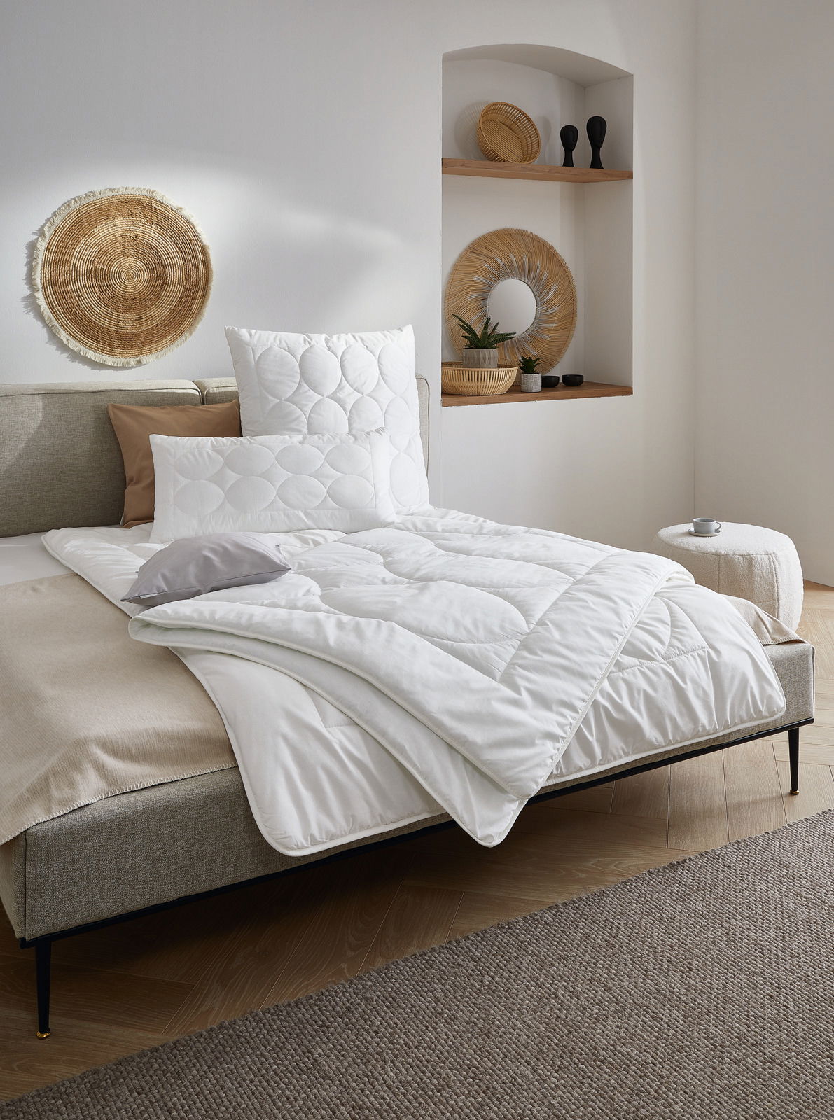 Gemütliches Schlafzimmer mit einem Bett, das mit einer weißen Bettdecke und Kissen ausgestattet ist. Die Perspektive zeigt das Bett von der Seite, mit dekorativen Elementen an der Wand und einem kleinen Beistelltisch.