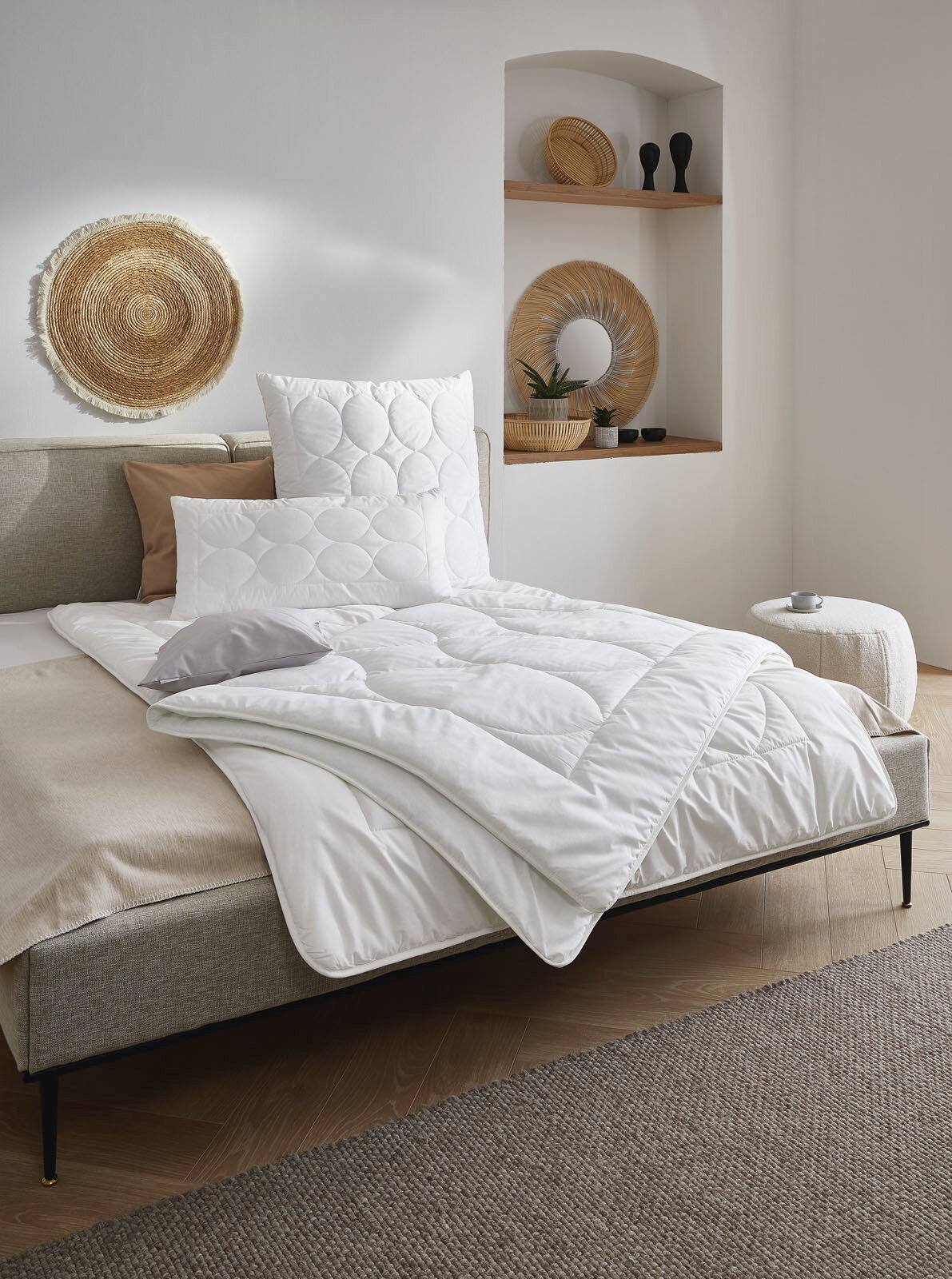 Gemütliches Schlafzimmer mit einem Bett, das mit einer weißen Bettdecke und Kissen ausgestattet ist. Die Perspektive zeigt das Bett von der Seite, mit dekorativen Elementen an der Wand und einem kleinen Beistelltisch.