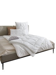 Gemütliches Schlafzimmer mit einem Bett, das mit einer weißen Bettdecke und Kissen ausgestattet ist. Die Perspektive zeigt das Bett von der Seite, mit dekorativen Elementen an der Wand und einem kleinen Beistelltisch.