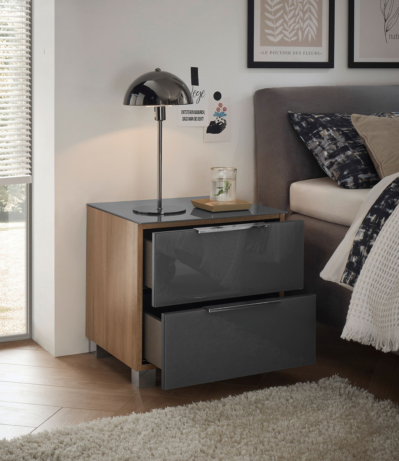 Moderne Nachtkommode mit zwei Schubladen in Hochglanzoptik, seitliche Holzverkleidung, abgebildet aus einer seitlichen Perspektive neben einem Bett mit Lampe und Dekoration.