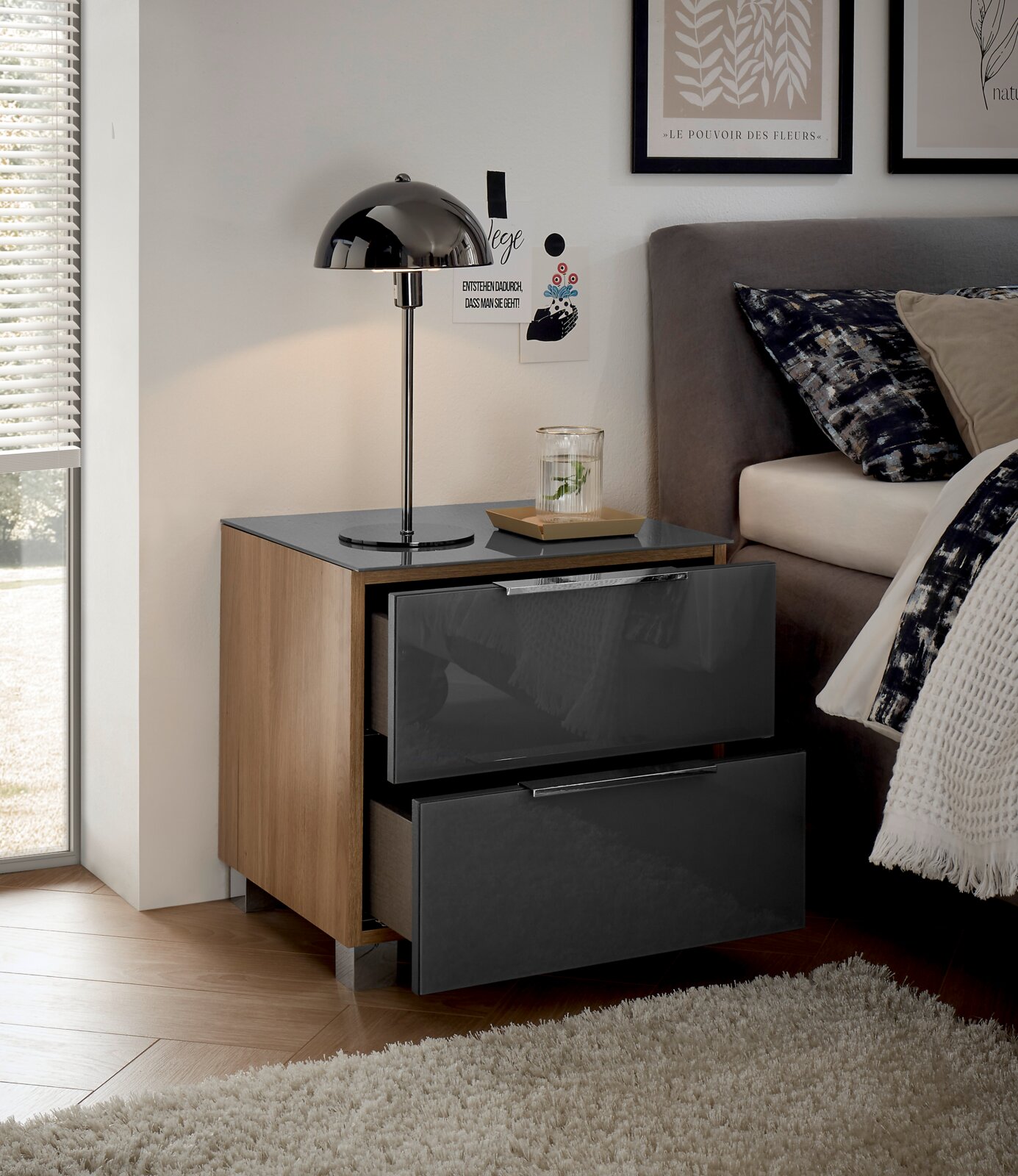 Moderne Nachtkommode mit zwei Schubladen in Hochglanzoptik, seitliche Holzverkleidung, abgebildet aus einer seitlichen Perspektive neben einem Bett mit Lampe und Dekoration.
