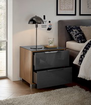 Moderne Nachtkommode mit zwei Schubladen in Hochglanzoptik, seitliche Holzverkleidung, abgebildet aus einer seitlichen Perspektive neben einem Bett mit Lampe und Dekoration.