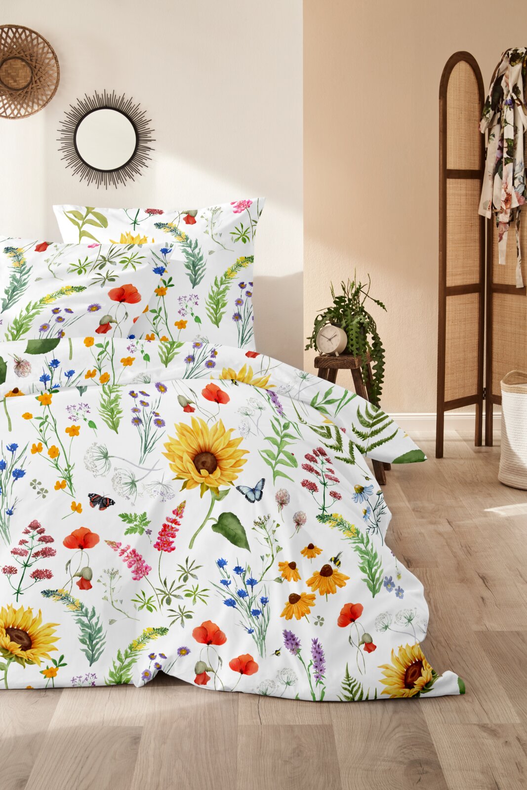 Bettwäsche-Set mit weißem Grund und buntem Wildblumen-Print (u. a. Sonnenblumen, Mohn, Wiesenblumen) auf Bett mit Kissen, fotografiert in leicht schräger Frontansicht im Schlafzimmer