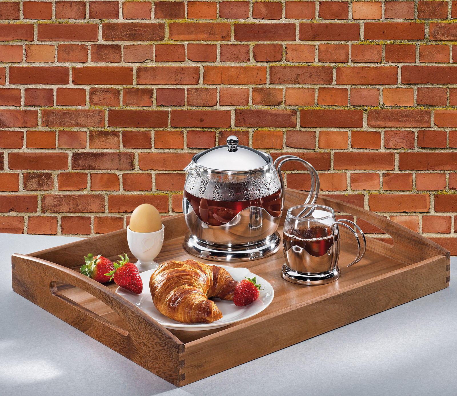 Holztablett mit Frühstücksset, bestehend aus Teekanne, Tasse, Croissant, Erdbeeren und Ei, von oben fotografiert.