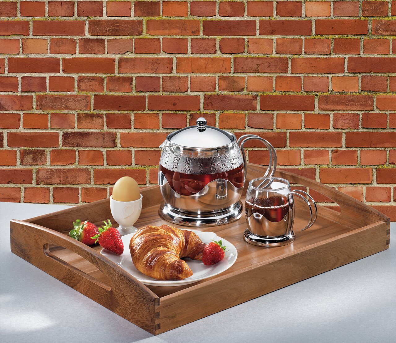 Houten dienblad met ontbijtset, bestaande uit een theepot, een kopje, een croissant, aardbeien en een ei, gefotografeerd van bovenaf.