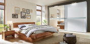 Houten nachtkastje met lade en opbergvlak, gezien vanaf de zijkant, naast een bed in een modern ingerichte slaapkamer.