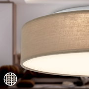 Detailaufnahme eines beigen Stoff-Lampenschirms einer Deckenleuchte mit leuchtender Unterseite, perspektivisch von unten/seitlich in einem Wohnraum