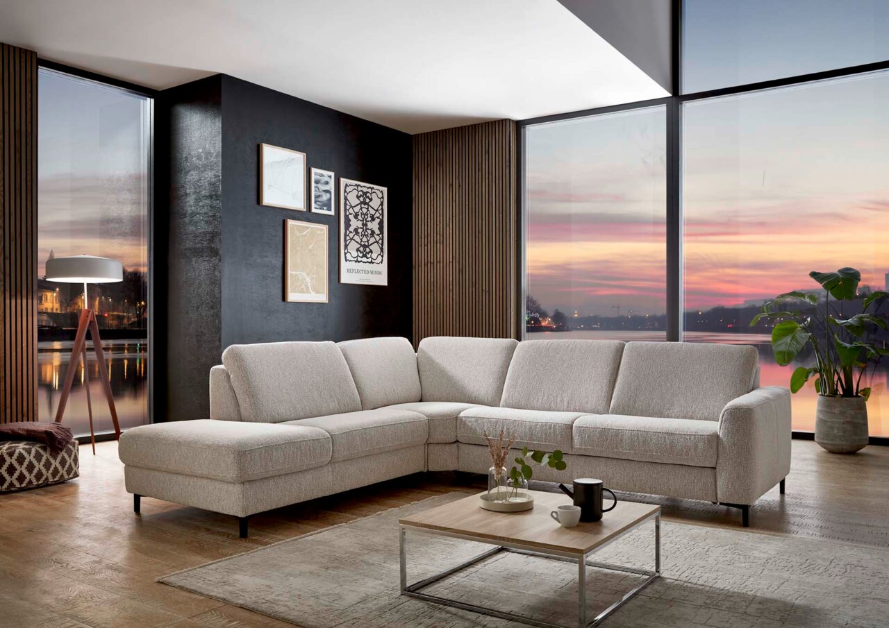 Valdera Ecksofa COMFORT VITAL Eckgarnitur in einem modernen Wohnzimmer mit Blick auf einen Sonnenuntergang durch große Fenster.