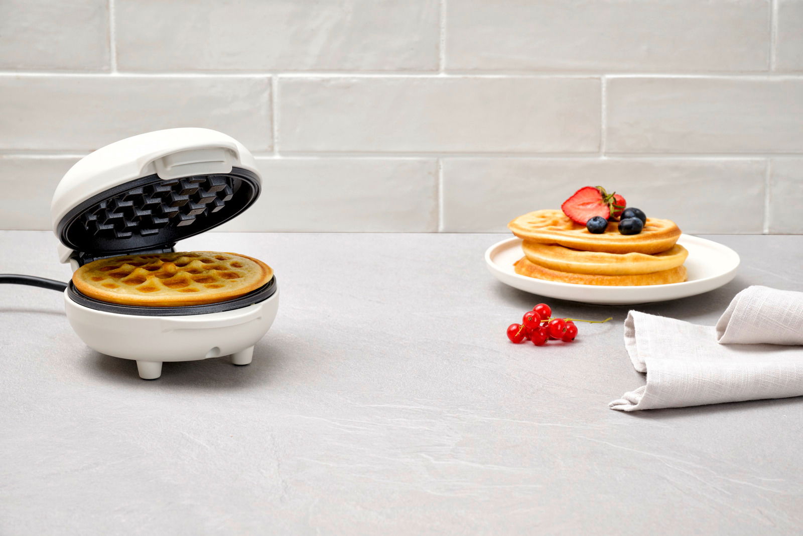 Ein weißes Waffeleisen mit einer frisch gebackenen Waffel, daneben ein Teller mit gestapelten Waffeln und frischen Beeren, aus einer seitlichen Perspektive fotografiert.