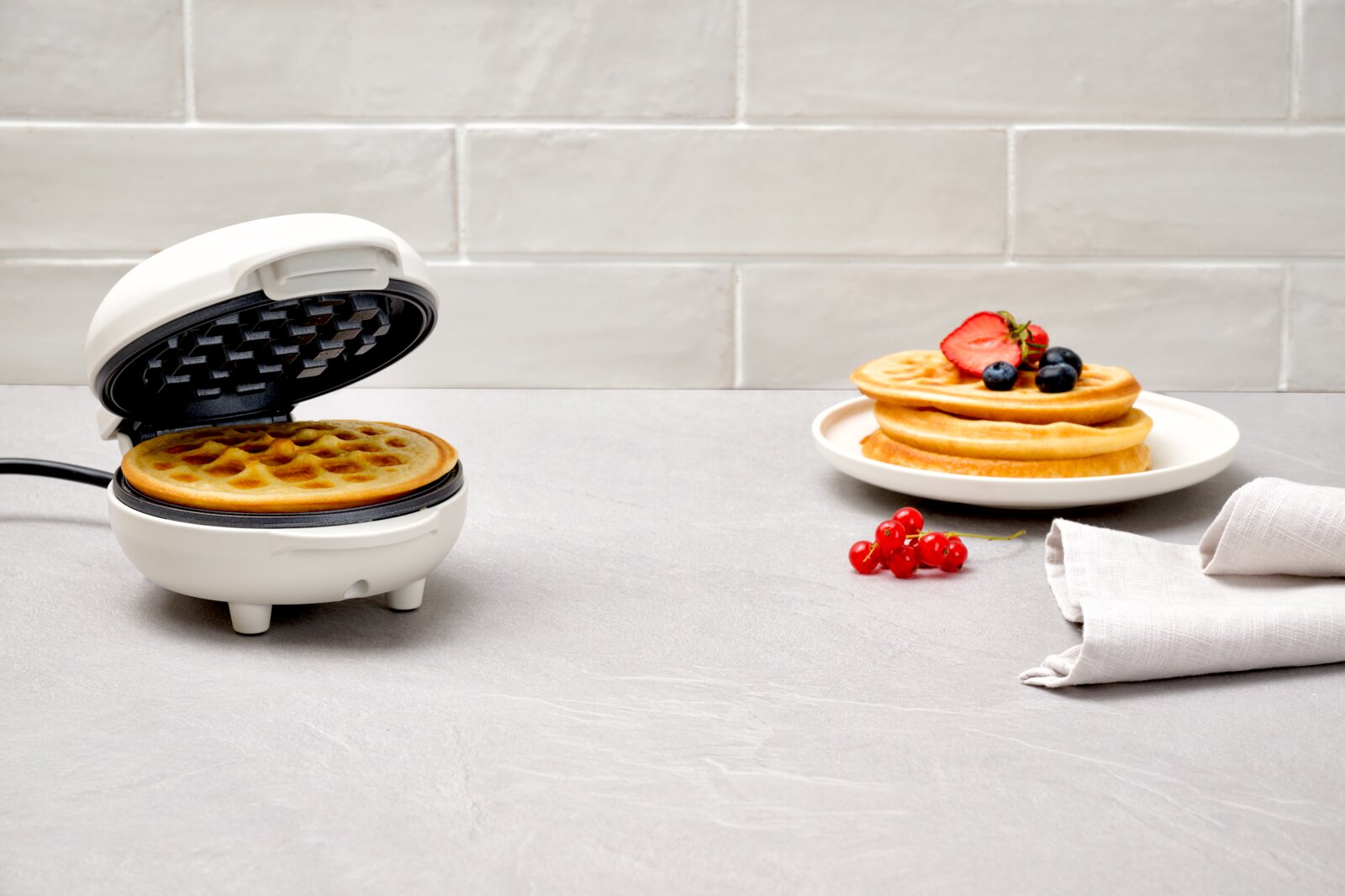 Ein weißes Waffeleisen mit einer frisch gebackenen Waffel, daneben ein Teller mit gestapelten Waffeln und frischen Beeren, aus einer seitlichen Perspektive fotografiert.