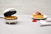 Ein weißes Waffeleisen mit einer frisch gebackenen Waffel, daneben ein Teller mit gestapelten Waffeln und frischen Beeren, aus einer seitlichen Perspektive fotografiert.