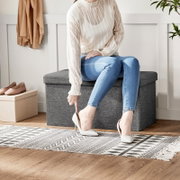 Frontale Ansicht: Person sitzt auf einer grauen Aufbewahrungsbank im Flur und zieht einen weißen High Heel an; daneben stehen beige Ballerinas und ein beiger Aufbewahrungskorb.