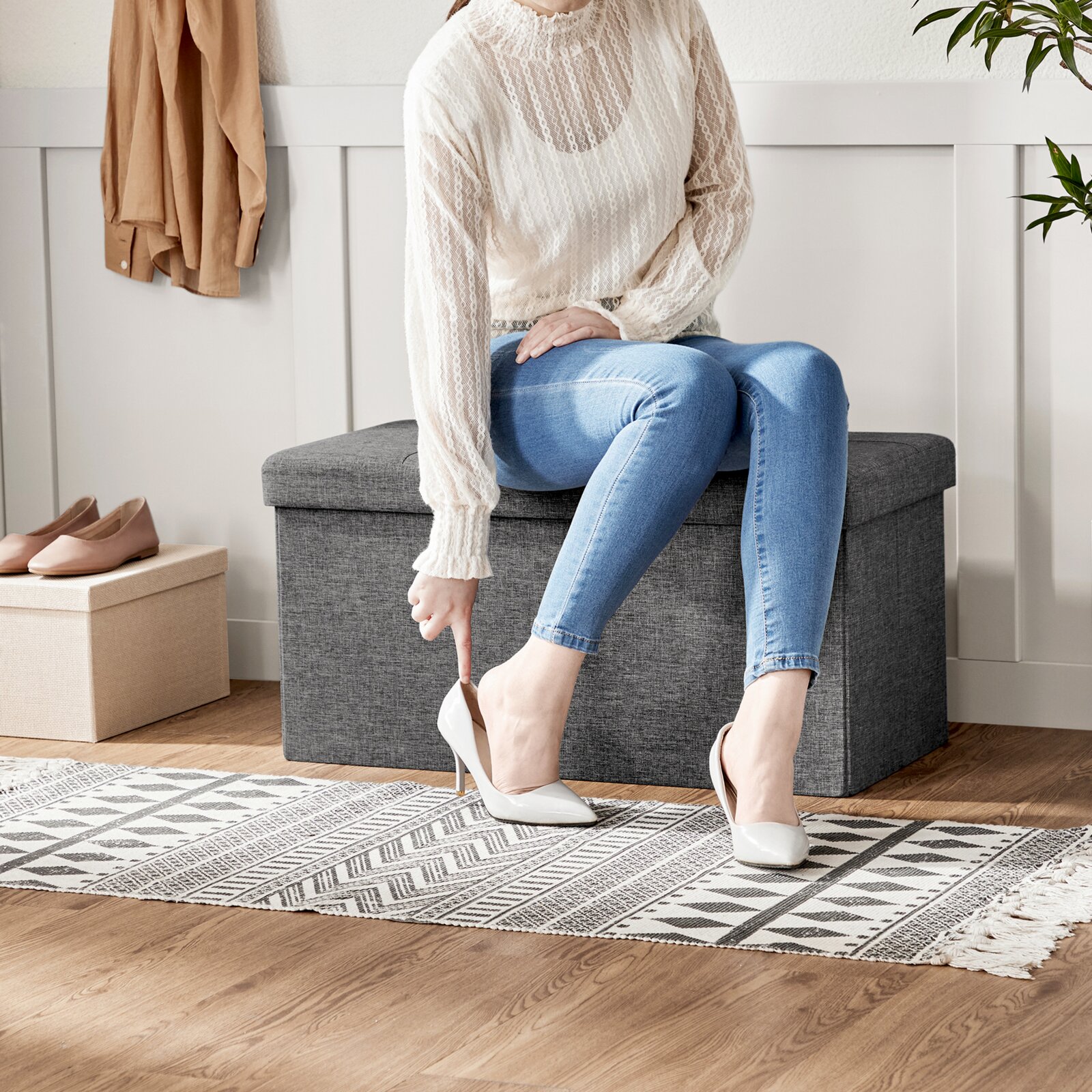 Frontale Ansicht: Person sitzt auf einer grauen Aufbewahrungsbank im Flur und zieht einen weißen High Heel an; daneben stehen beige Ballerinas und ein beiger Aufbewahrungskorb.