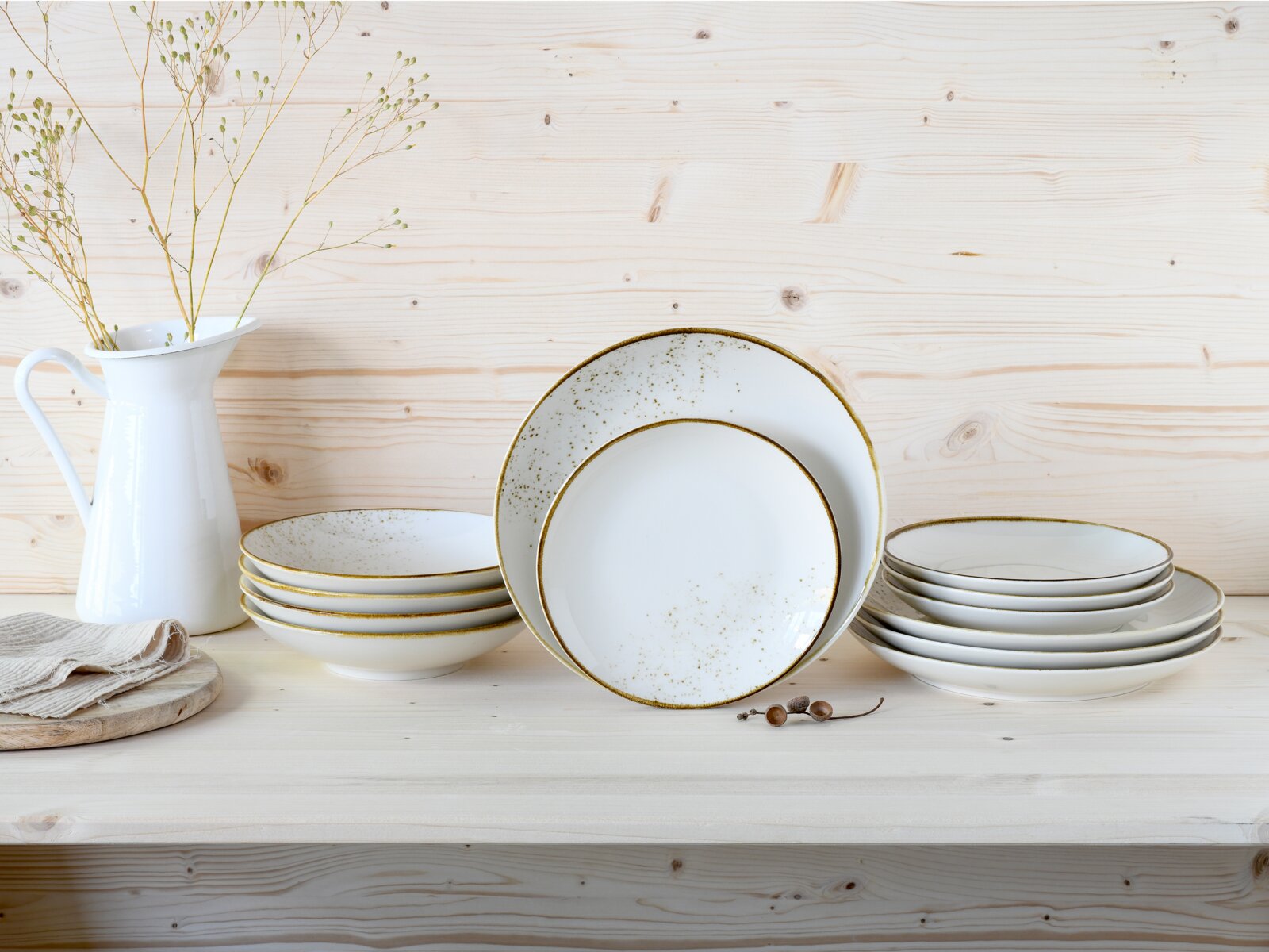 Creatable Tellerset 12-tlg NATURE COLLECTION Weißes Keramik-Geschirrset mit goldfarbenem Rand und feiner Sprenkel-Optik, gestapelte Teller und Schalen auf hellem Holztisch neben weißer Vase, frontal in leichter Draufsicht fotografiert