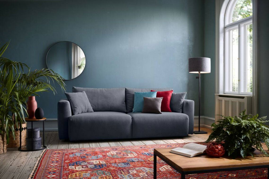 Graues Funktionssofa in einem stilvoll eingerichteten Wohnzimmer mit blauer Wand, rundem Spiegel, Stehlampe und buntem Teppich, Perspektive von vorne.