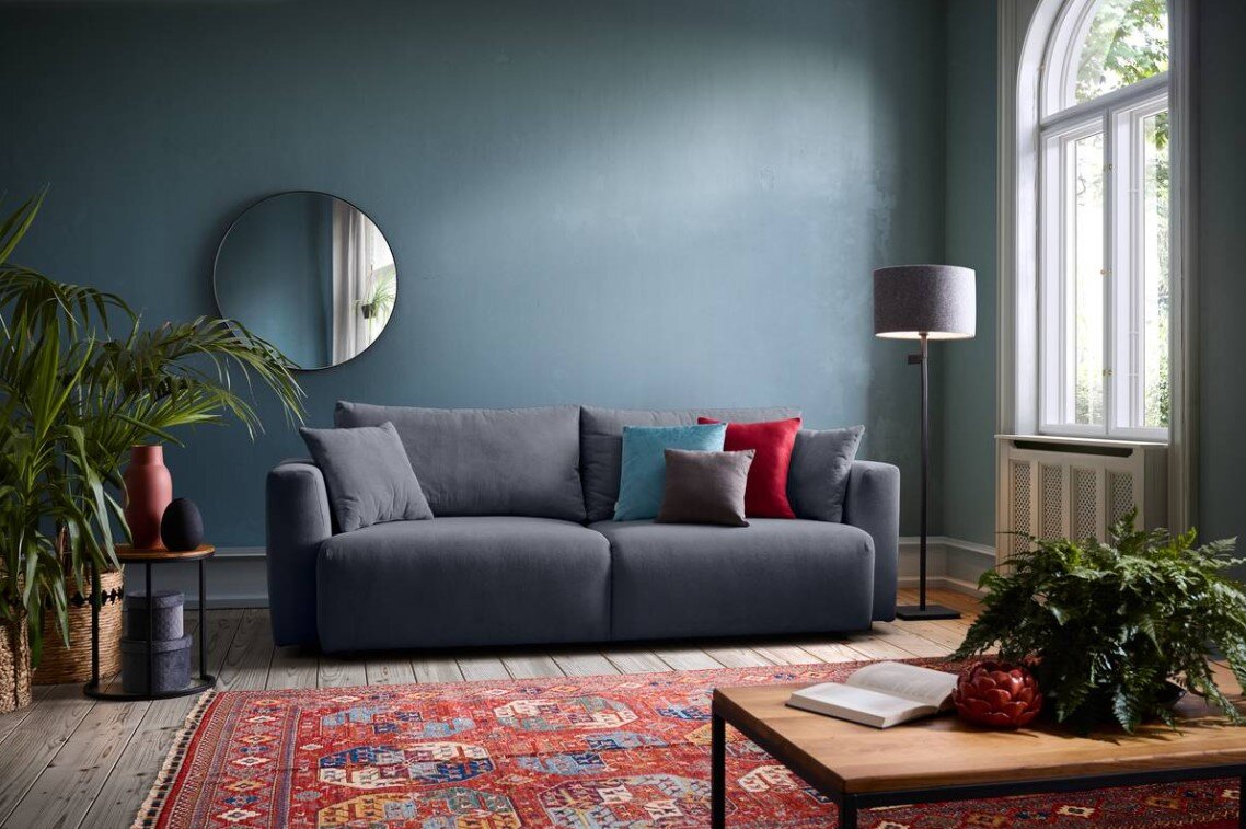 Graues Funktionssofa in einem stilvoll eingerichteten Wohnzimmer mit blauer Wand, rundem Spiegel, Stehlampe und buntem Teppich, Perspektive von vorne.