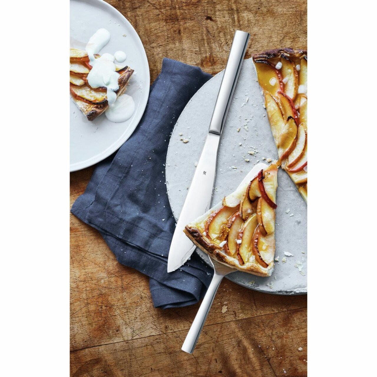 WMF Taartbestekset, 2-delig NUOVA Taartset Nuova met een elegant mes en een taartschep, gepresenteerd op een houten tafel met stukjes appeltaart, gezien van bovenaf.