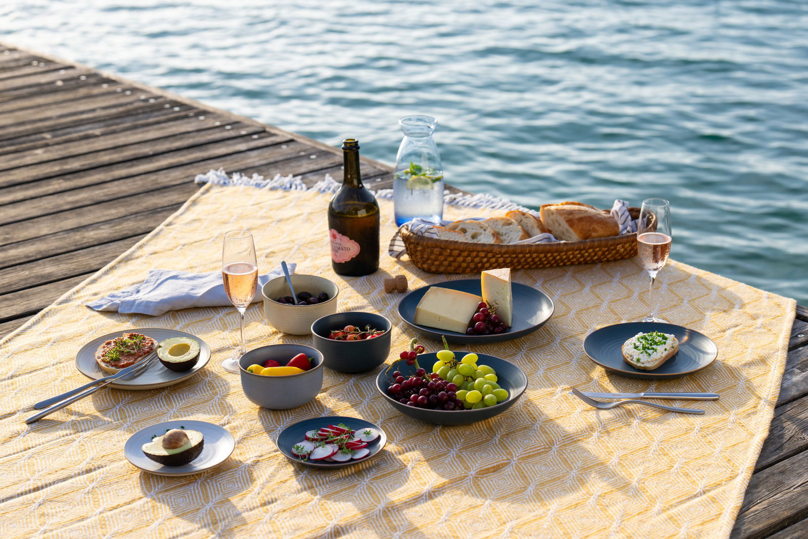 Frühstücksteller 22 cm auf einem Picknick am Seeufer, gefüllt mit Käse, Trauben und Brot, umgeben von weiteren Speisen und Getränken auf einer gelben Decke.