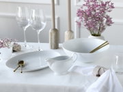 Elegantes weißes Porzellan-Set mit geriffelter Schale, Teller und Sauciere auf gedecktem Tisch, dazu goldfarbenes Besteck, Weingläser und violette Blumen; Perspektive: leicht erhöhte Frontansicht