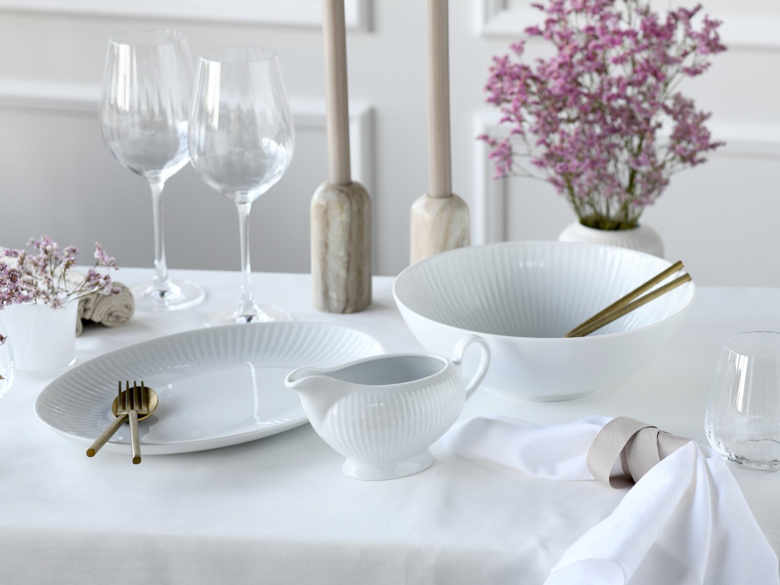 Elegantes weißes Porzellan-Set mit geriffelter Schale, Teller und Sauciere auf gedecktem Tisch, dazu goldfarbenes Besteck, Weingläser und violette Blumen; Perspektive: leicht erhöhte Frontansicht