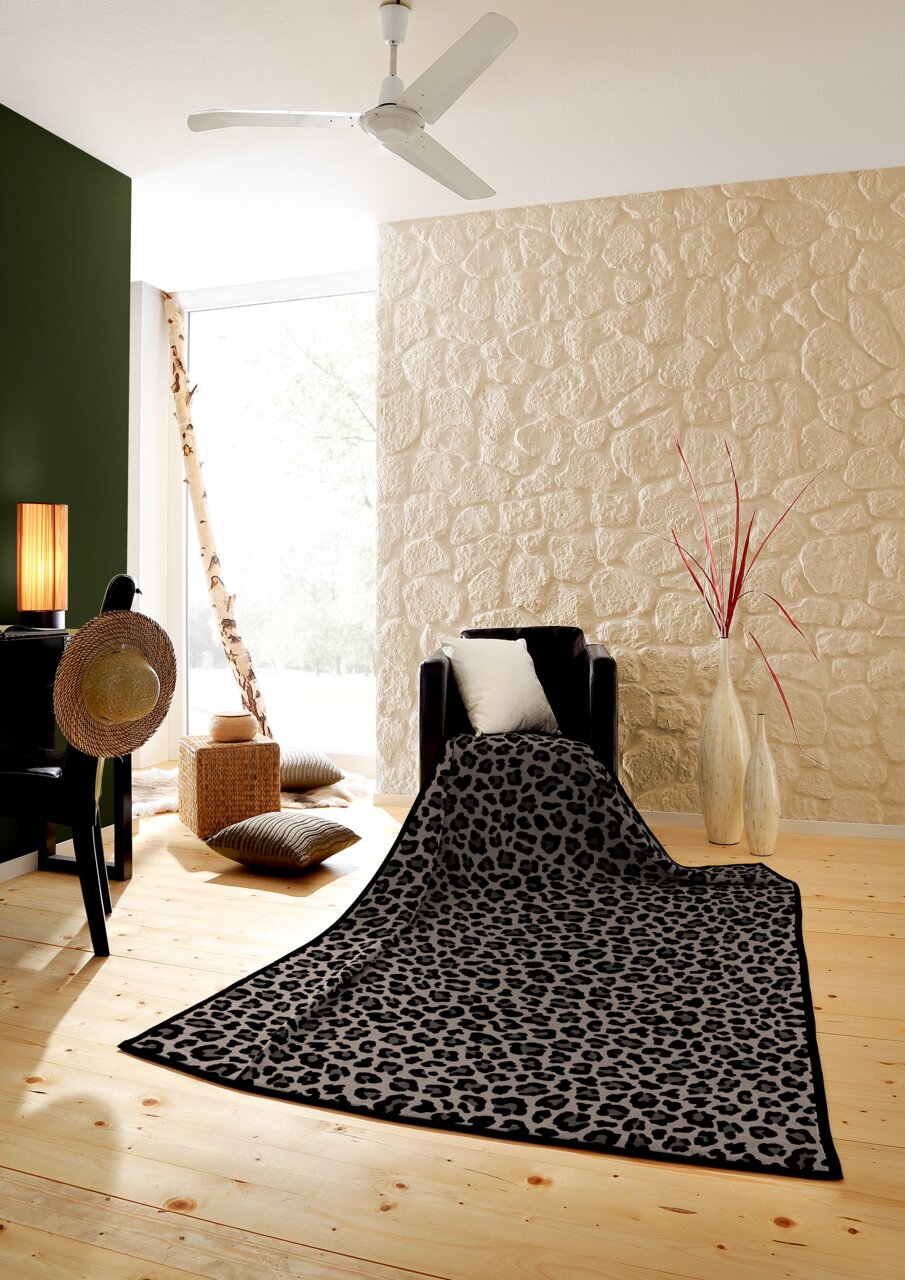 Leopardengemusterte Wohndecke auf einem Sessel in einem stilvoll eingerichteten Raum mit Holzboden und Steinwand, aus der Perspektive von vorne.