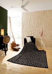 Leopardengemusterte Wohndecke auf einem Sessel in einem stilvoll eingerichteten Raum mit Holzboden und Steinwand, aus der Perspektive von vorne.